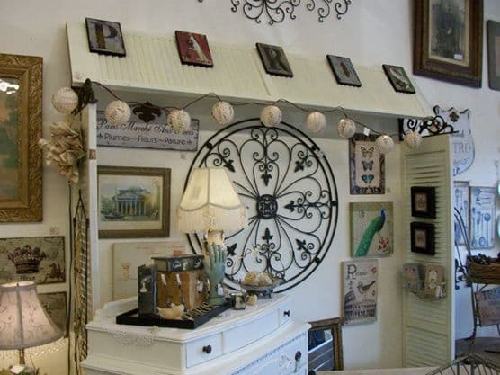 Wall display with French-inspired signs, an ornate iron medallion, a white dresser with a lamp, and paper lanterns